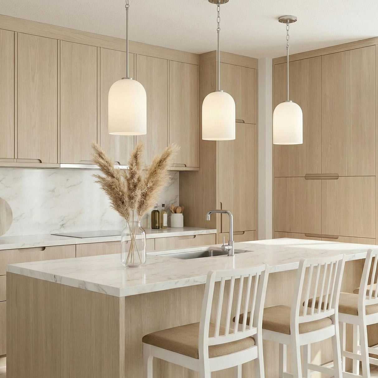 A minimalist kitchen featuring three Elira Pendant 9", Brushed Nickel fixtures with frosted glass shades installed over a white marble island surrounded by light wood cabinetry.