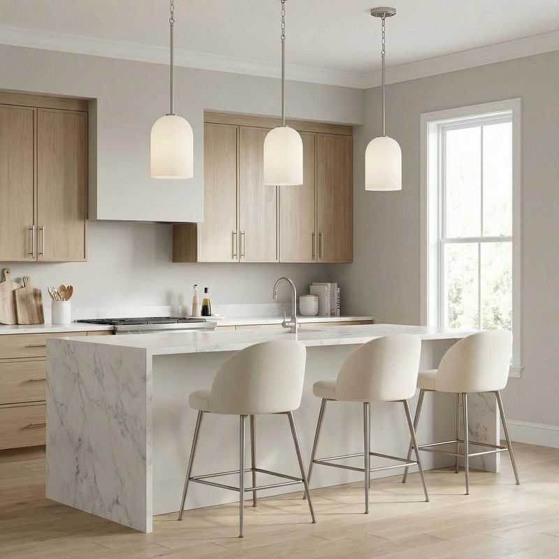 A contemporary kitchen featuring three Elira Pendant 9", Brushed Nickel fixtures with frosted glass shades over a white marble waterfall island and light wood cabinets.