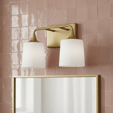 Harper 2 Light Vanity, Aged Brass - A dual-bulb vanity light with white frosted shades and an aged brass finish is mounted on a pink tiled wall above a gold-framed mirror.