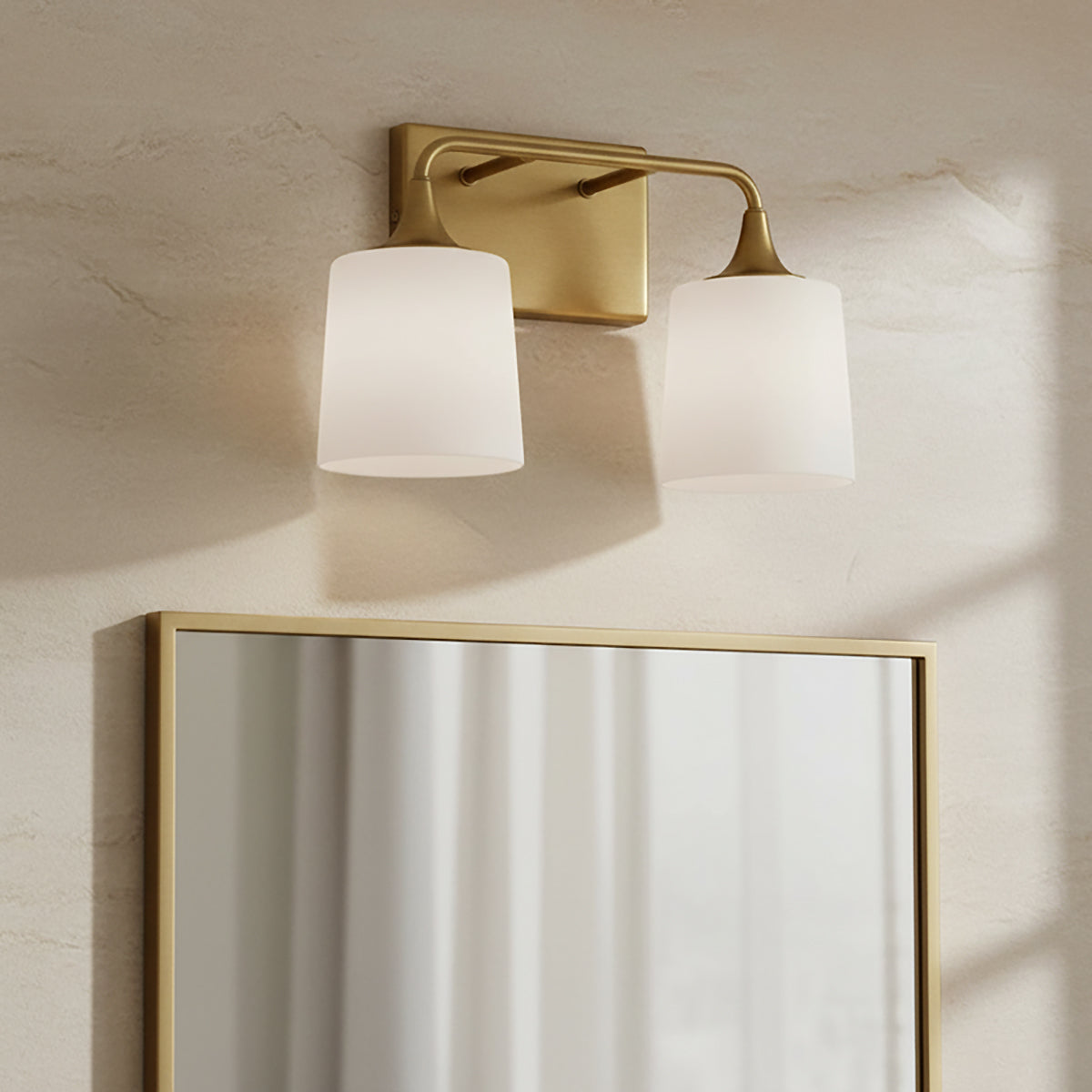 Harper 2 Light Vanity, Aged Brass - A brass vanity light with two frosted glass shades mounted on a textured wall above a brass-framed mirror.