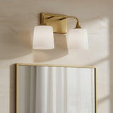 Harper 2 Light Vanity, Aged Brass - A brass vanity light with two frosted glass shades mounted on a textured wall above a brass-framed mirror.