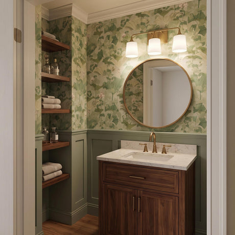 A traditional bathroom featuring the Harper 3 Light Vanity, Aged Brass with white shades above a round mirror, green botanical wallpaper, and a walnut vanity cabinet.