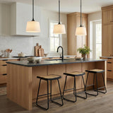 A transitional kitchen features three matte black pendants with white glass shades over a wood island with a dark soapstone countertop and three rattan stools.