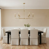 Harper 6 Light Chandelier 49, Aged Brass - A linear aged brass chandelier hangs above a dark wood dining table with upholstered chairs.