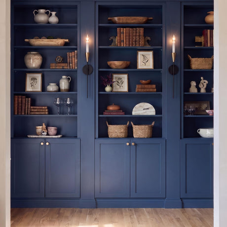 A transitional living room with the Astra Wall Sconce 22", Aged Brass and Black, mounted on navy blue built-in bookshelves featuring books and decorative vases.