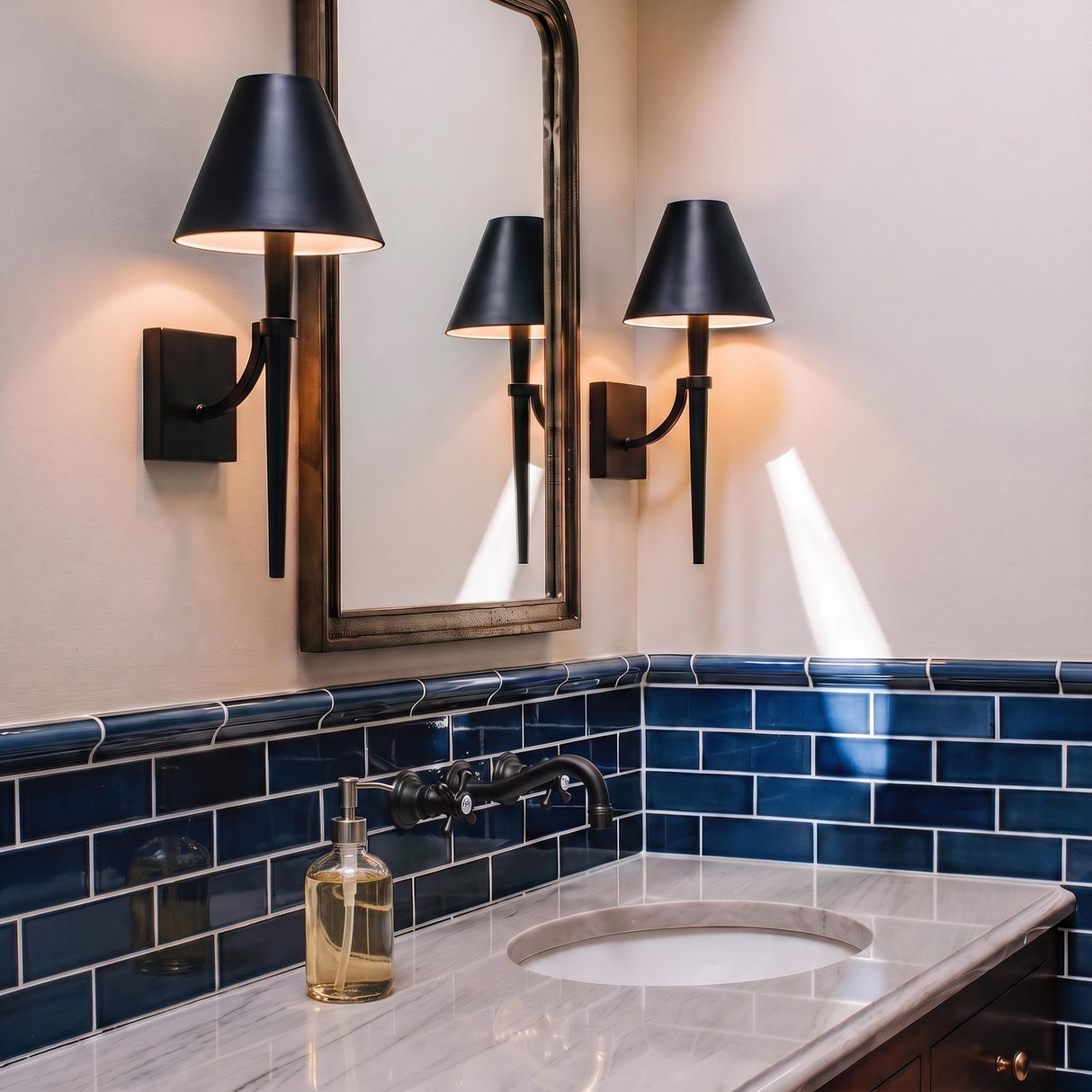 A transitional bathroom with two matte black Clara Wall Sconce 19" fixtures flanking a bronze-framed mirror above a white marble countertop and blue subway tile.