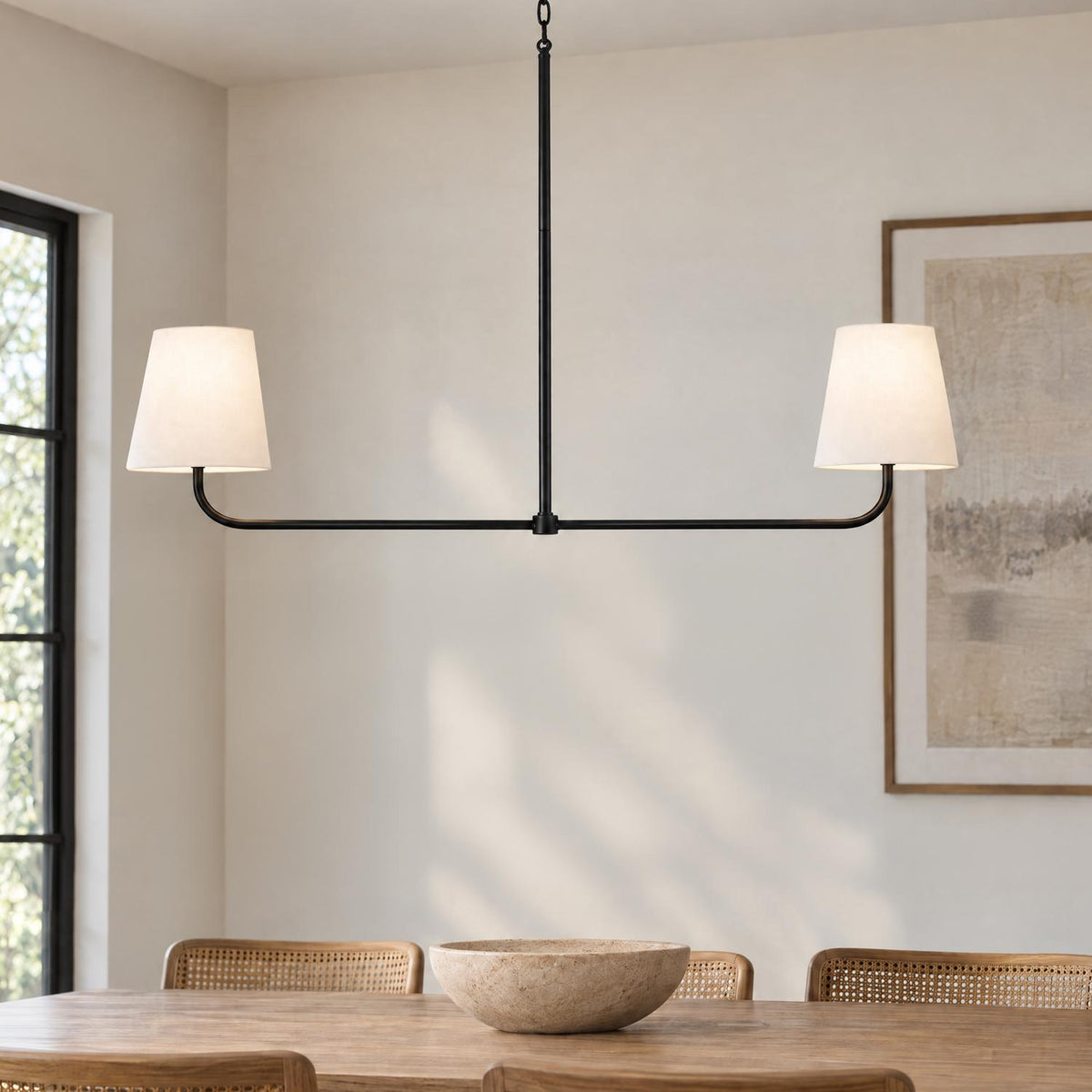 A transitional dining room with a matte black linear pendant featuring off-white linen shades hanging over a natural wood dining table and cane back chairs.