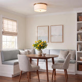 A transitional dining room with the Wells Flush Mount 16", Aged Brass with a white linen shade mounted over a round wood table and light blue banquette.