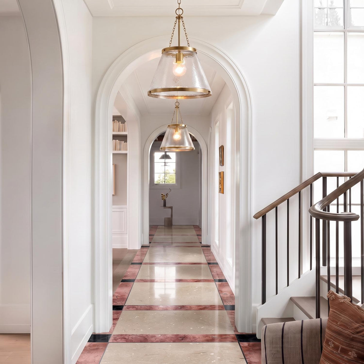 A transitional foyer with two Elora Pendant 15", Aged Brass with Hammered Glass fixtures along a ceiling of arched hallways with inlaid stone tile flooring and white walls.