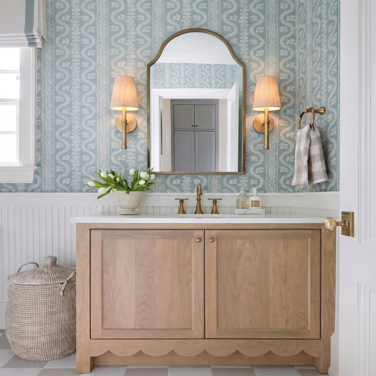 A cottage style bathroom with two matte brass wall sconces with linen shades flanking an arched mirror over a scalloped wood vanity and patterned blue wallpaper.