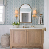 A cottage style bathroom with two matte brass wall sconces with linen shades flanking an arched mirror over a scalloped wood vanity and patterned blue wallpaper.