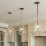 A traditional kitchen features three Delmar Pendant 11", Aged Brass fixtures with aged brass chains and faceted glass shades over the island alongside cream cabinetry.