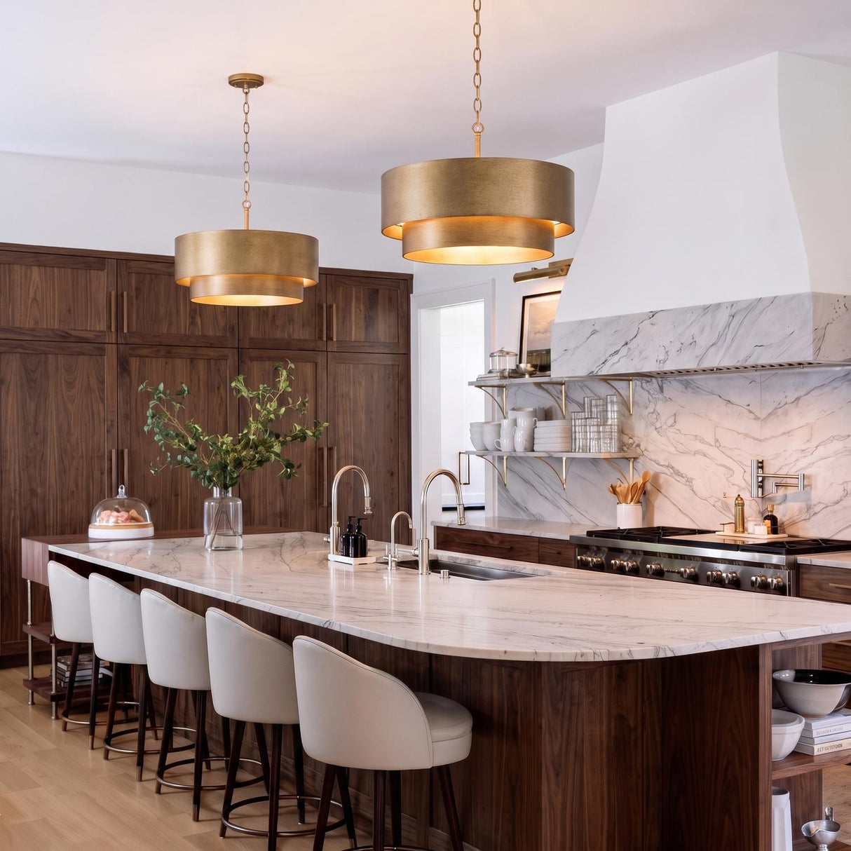A transitional kitchen with two brushed brass tiered drum pendants over a white marble island featuring walnut cabinetry and four cream leather barstools.