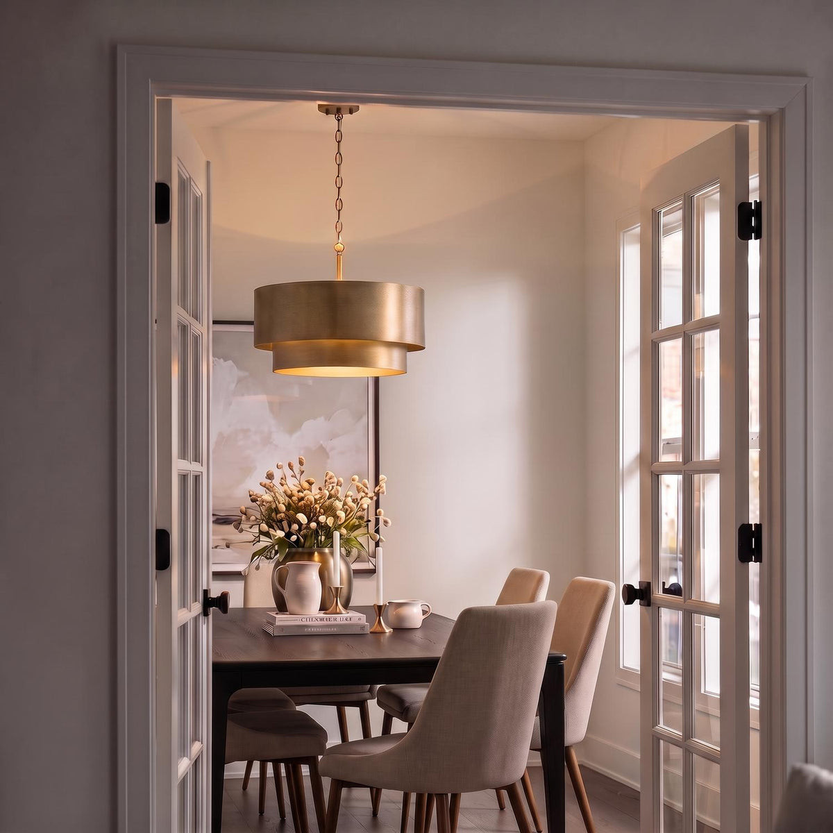A transitional dining room framed by open French doors features a textured brushed brass pendant light hanging above a dark wooden dining table set with upholstered chairs.