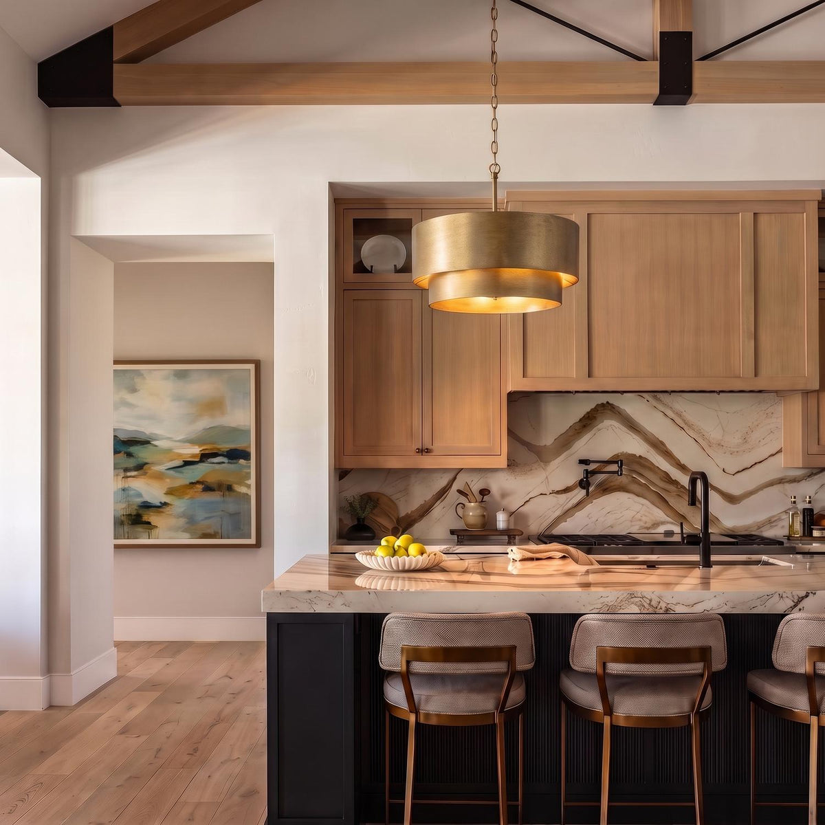 A transitional kitchen features a brushed brass tiered drum pendant suspended over a veined marble island surrounded by light wood cabinetry and exposed ceiling beams.