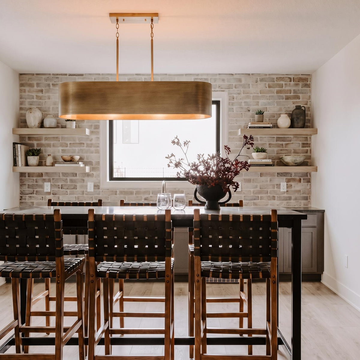 A modern farmhouse dining room with a brushed brass linear pendant over a black high-top table, featuring a white-washed brick wall and floating shelves.