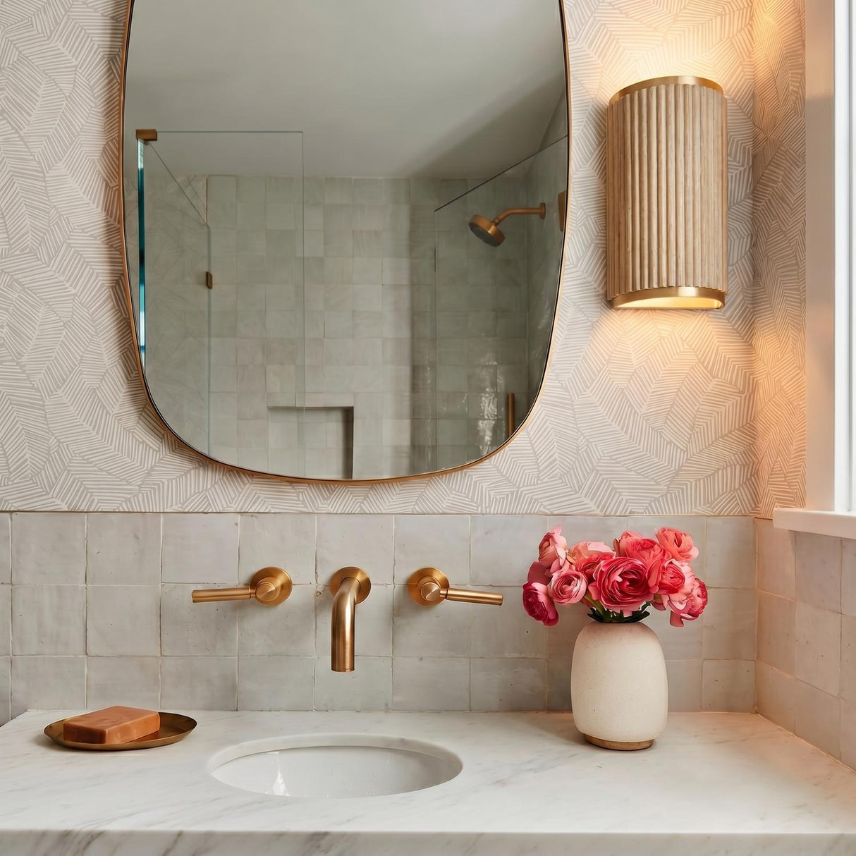 A transitional bathroom featuring the Leery Wall Sconce 12-inch in whitewash and matte brass beside a brass-rimmed mirror above a marble countertop and white Zellige tile backsplash.