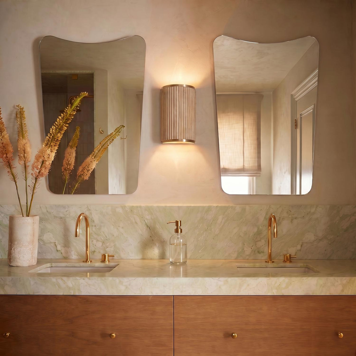 A mid-century bathroom with the ribbed whitewash and matte brass Leery Wall Sconce 12 inch centered between asymmetrical mirrors above a green marble vanity and wood cabinets.