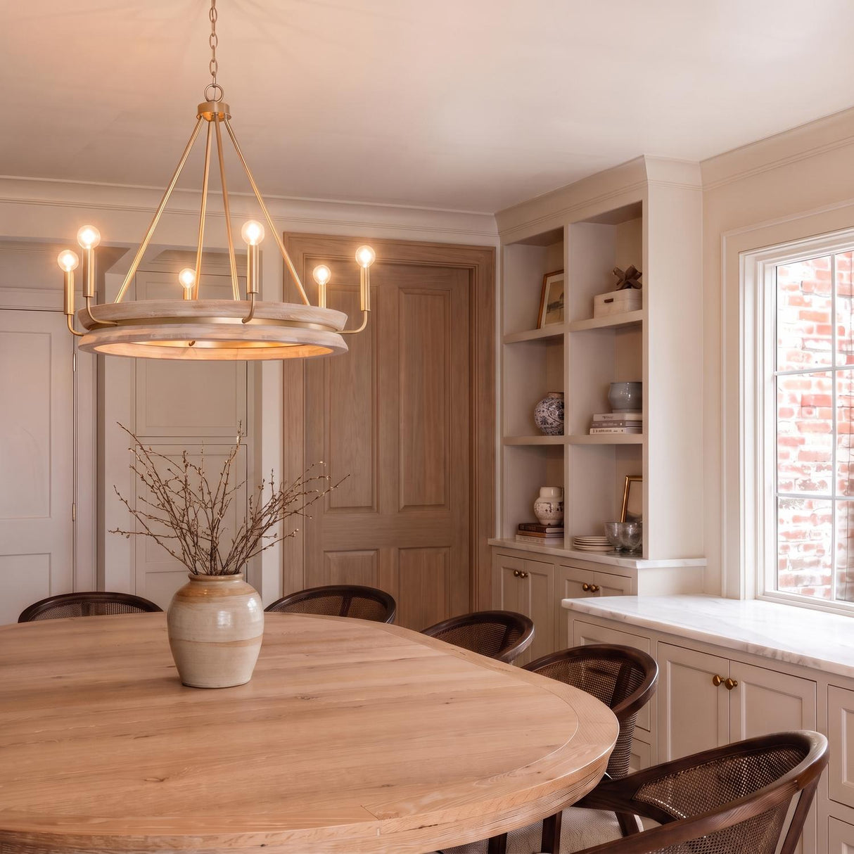 A transitional dining room featuring the Maren 6 Light Chandelier in whitewash and matte brass over a round wood table with cane-back chairs and built-in cabinets.
