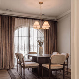 A transitional dining room with the Simone 4 Light Chandelier in matte brass and linen shades over a dark wood table with brown velvet curtains.