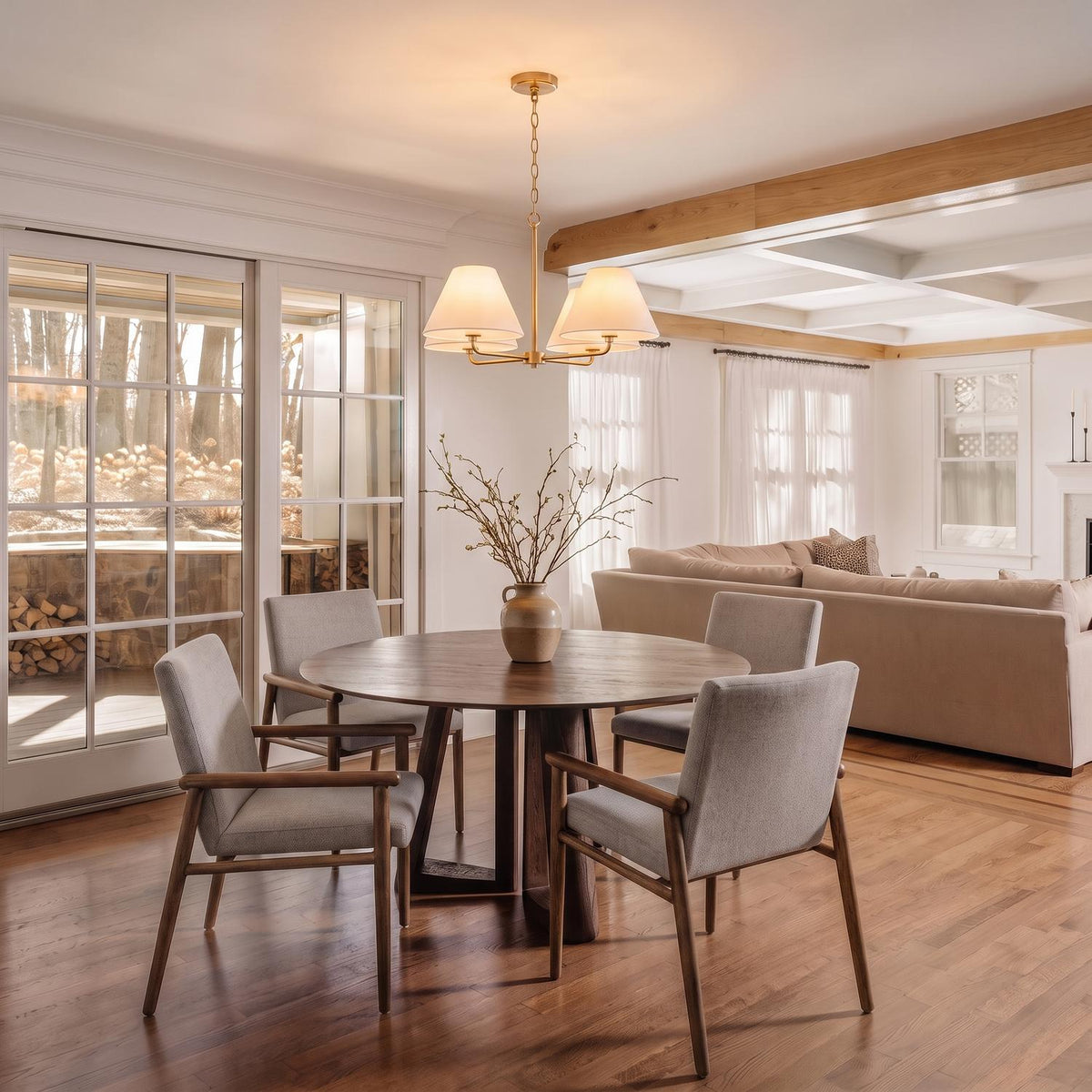 A transitional dining room with the Simone 4 Light Chandelier 27", Matte Brass over a round walnut table and grey upholstered chairs near large glass doors.