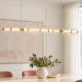 A contemporary dining room featuring the Elara LED 9 Light Linear Chandelier in brass and clear glass suspended over a marble dining table with pink chairs.