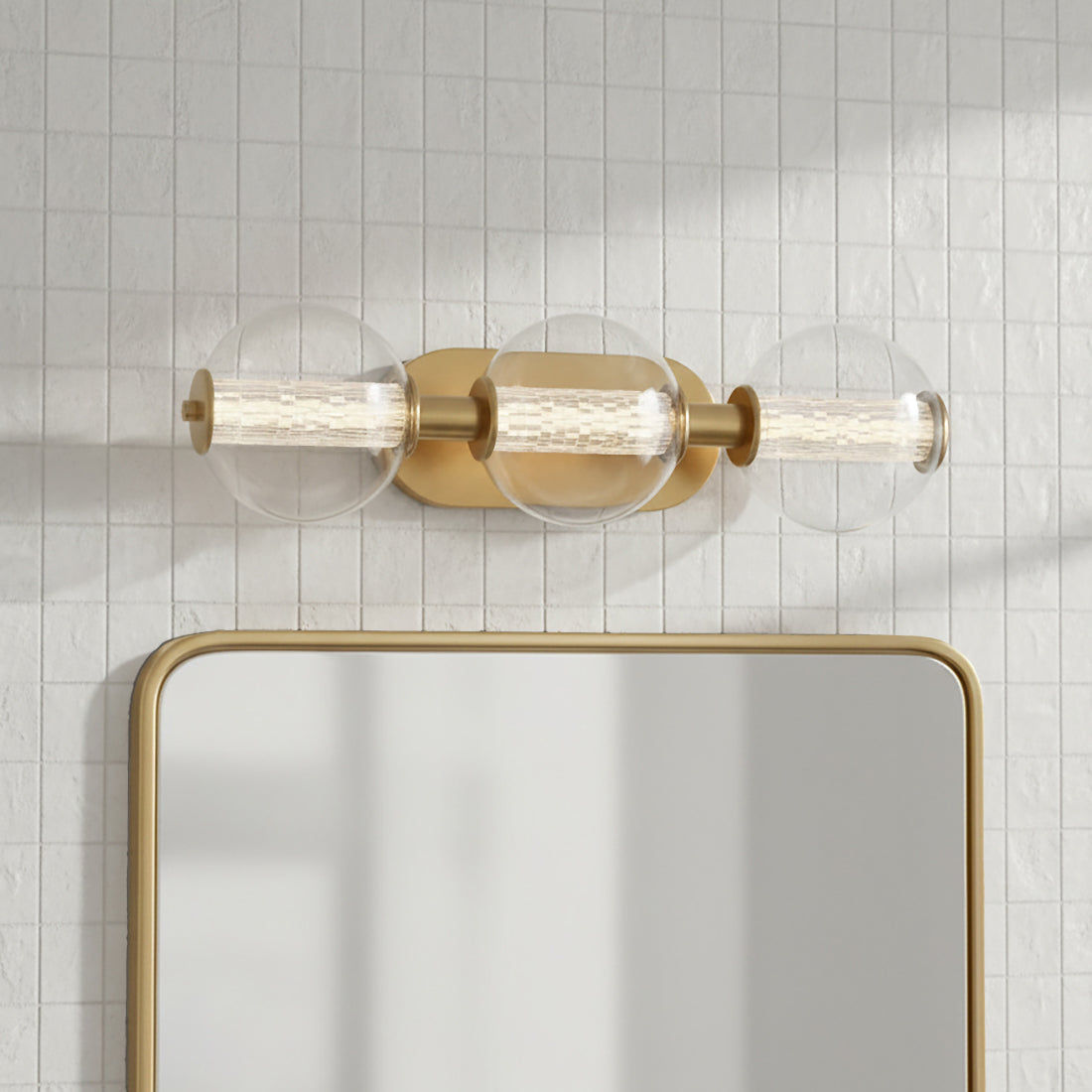 Elara LED 3 Light Vanity, Brass and Clear Glass - A brass vanity light with three clear glass globes containing ribbed glass cylinders is mounted above a mirror against a tiled wall.