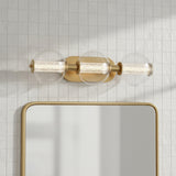 Elara LED 3 Light Vanity, Brass and Clear Glass - A brass vanity light with three clear glass globes containing ribbed glass cylinders is mounted above a mirror against a tiled wall.