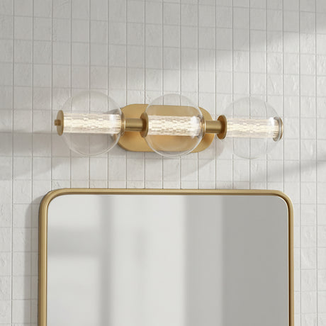 Elara LED 3 Light Vanity, Brass and Clear Glass - A brass vanity light with three clear glass globes containing ribbed glass cylinders is mounted above a mirror against a tiled wall.