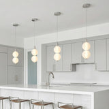 A minimalist white kitchen features four polished nickel pendants with ribbed opal glass globes over the white quartz island, gray flat-panel cabinetry, and silver bar stools.