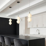 A minimalist kitchen features three Nola LED 2 Light Pendant 6", Polished Nickel and Ribbed Opal Glass fixtures over a black island under a white beamed ceiling.
