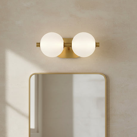 Nola LED 2 Light Vanity 15, Brass and Ribbed Opal Glass - A brass vanity light with two ribbed opal glass globes mounted above a gold-framed mirror against a textured cream wall.