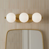 Nola LED 3 Light Vanity 23, Brass and Ribbed Opal Glass - A brass vanity light with three ribbed opal glass globes above a rounded gold mirror, set against a backdrop of vertically stacked beige tiles.