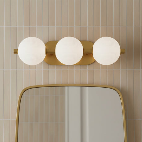 Nola LED 3 Light Vanity 23, Brass and Ribbed Opal Glass - A brass vanity light with three ribbed opal glass globes above a rounded gold mirror, set against a backdrop of vertically stacked beige tiles.