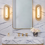 A contemporary bathroom features Vance LED Wall Sconce 16", Brass and Textured Glass fixtures flanking a brass mirror above a white marble vanity.