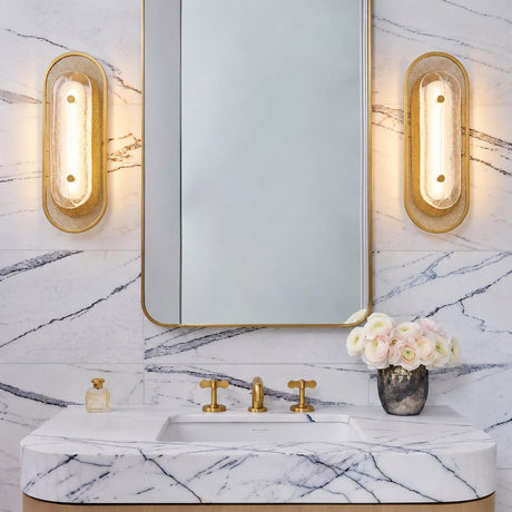 A contemporary bathroom features Vance LED Wall Sconce 16", Brass and Textured Glass fixtures flanking a brass mirror above a white marble vanity.