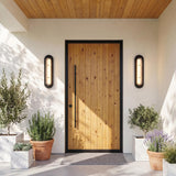 A modern home entry with two matte black Vance LED Outdoor Wall Lights flanking a light wood door on white stucco, with potted lavender and rosemary.