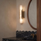 A contemporary bathroom featuring a Delya LED Wall Sconce in antique brass and alabaster mounted beside a round wood mirror above a scalloped marble backsplash.