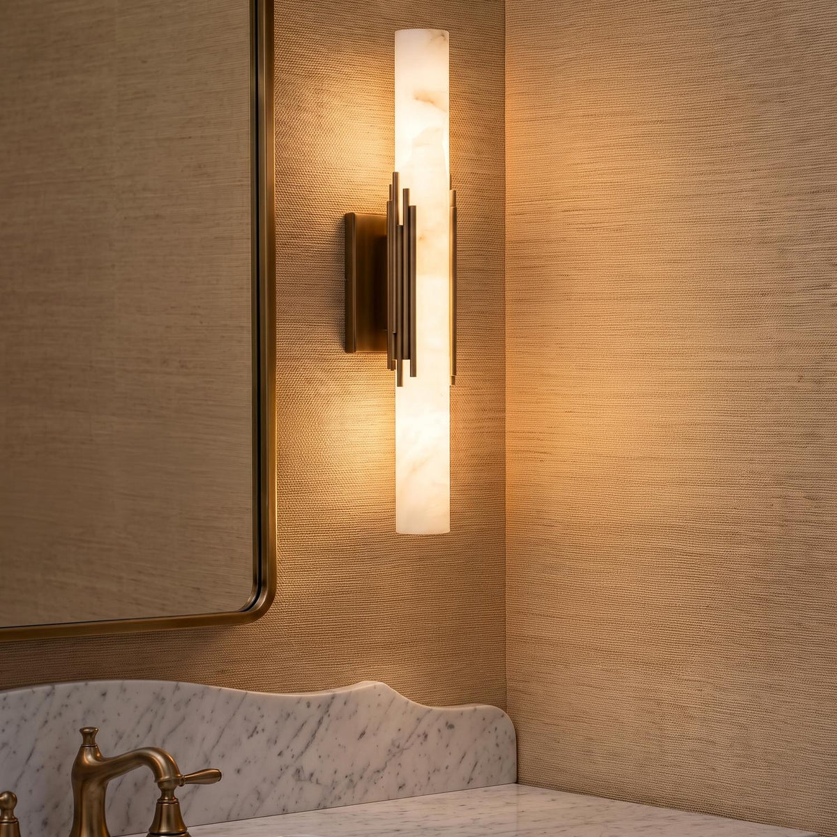 A contemporary bathroom with the Delya LED Wall Sconce 22" in antique brass and alabaster mounted beside a brass-framed mirror over a white marble vanity.