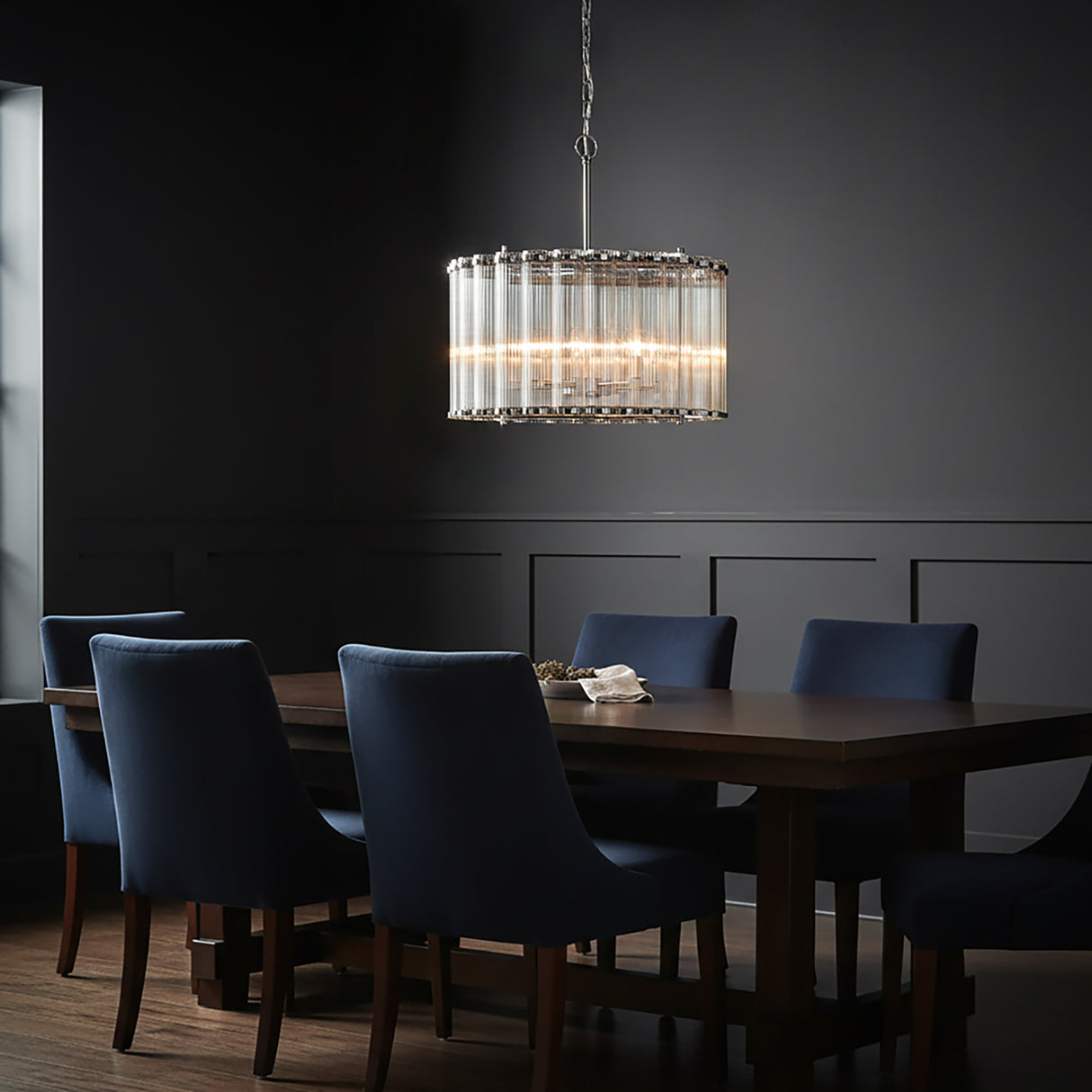 Moody dining room with the Nori 6 Light Chandelier 24", Polished Nickel and Ribbed Glass finish, hanging above a wooden table and navy blue chairs.