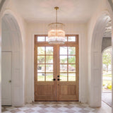 A transitional foyer with a brushed brass and ribbed glass chandelier centered over a checkered marble floor and light wood double front doors with white paneled arches.