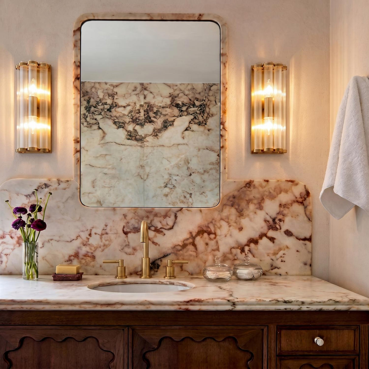 A contemporary bathroom featuring two Nori Wall Sconce 14 inch Brass and Ribbed Glass fixtures flanking a rounded mirror above a purple-veined marble countertop and backsplash.