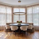 A contemporary dining room featuring the Lune LED Pendant 48", Brass and Black over a dark round dining table with beige chairs and sheer drapes.