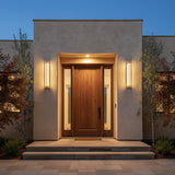 Prescott LED Outdoor Wall Light 24, Gold and Clear Glass - Two contemporary wall sconces flank a solid wood front door with glass sidelights, illuminating the entryway at dusk.