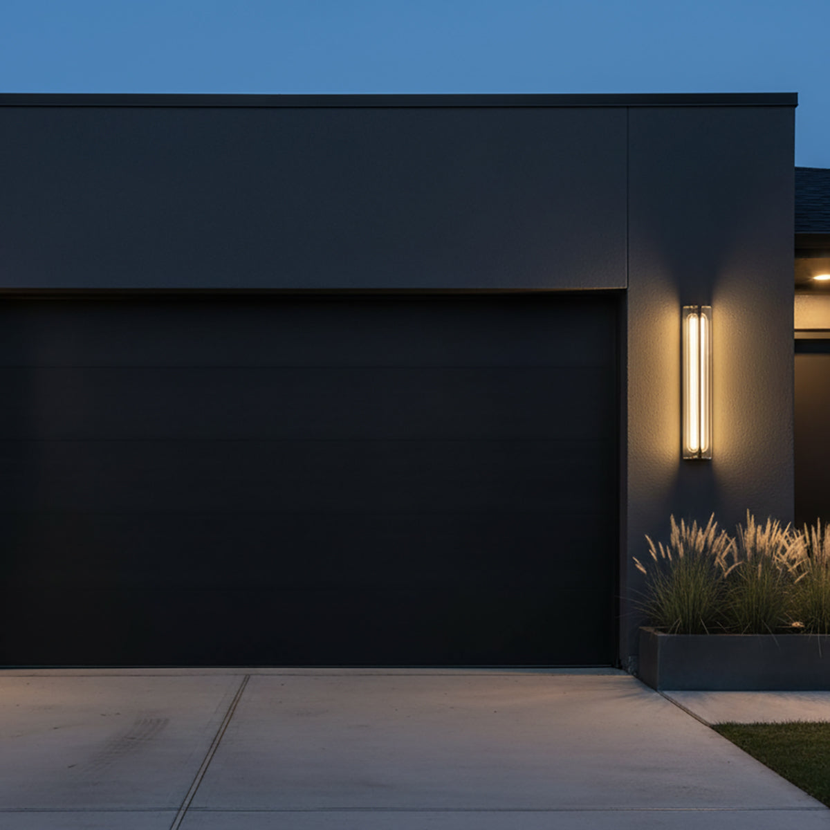 Prescott LED Outdoor Wall Light 24 - A contemporary outdoor wall light with a matte black finish and clear glass, mounted on a dark gray exterior wall next to a large black garage door, illuminating decorative grasses in a planter box.