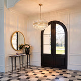 A transitional foyer with the Maxwell 12 Light Chandelier in Crystal and Aged Brass hanging over patterned marble tile near a black arched double door and white paneling.