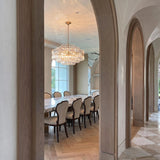 A Mediterranean dining room with the Maxwell 16 Light Chandelier 28", Crystal with Aged Brass hanging over a marble table between wooden arched doorways.