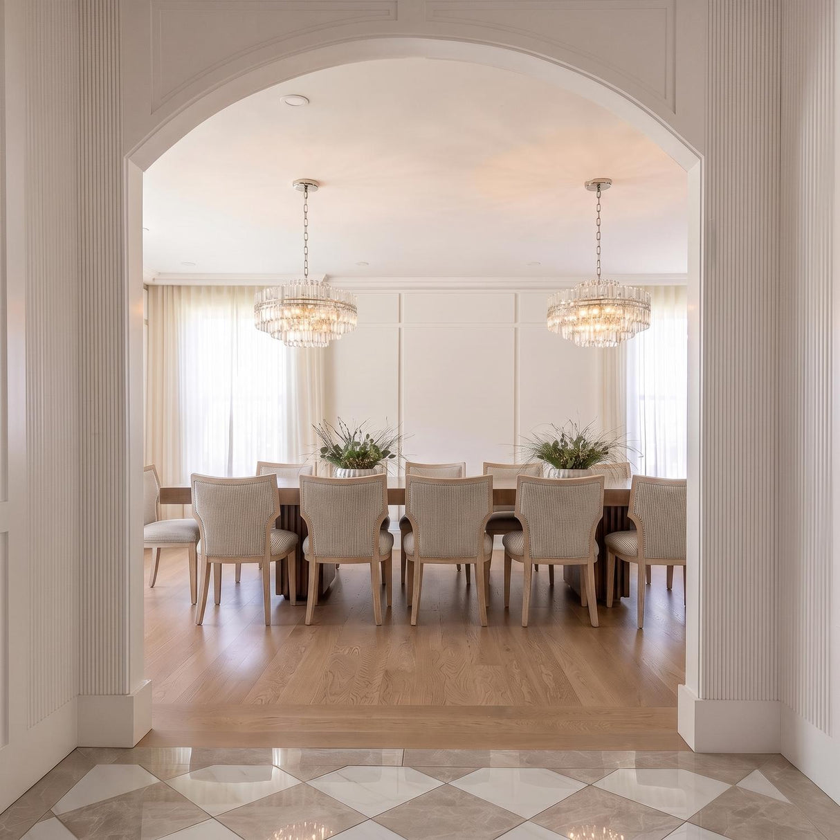 A transitional dining room featuring two Maxwell 16 Light Chandelier 28-inch crystal and polished nickel fixtures hanging over a wood table viewed through a white arched entryway.