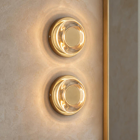 A modern bathroom with two Cam LED Wall Sconces in polished brass and clear glass mounted vertically on a textured beige plaster wall beside a slim brass mirror.