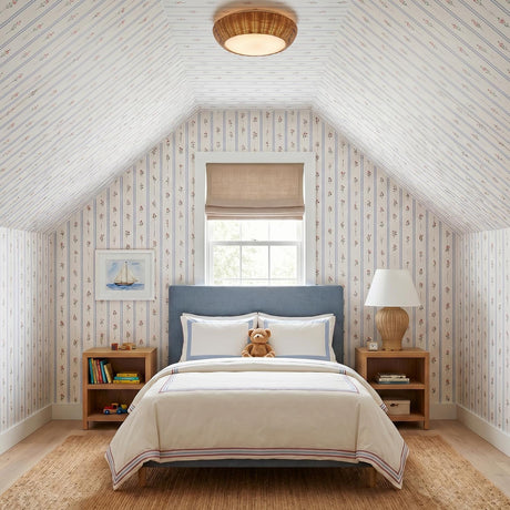 A coastal bedroom featuring the Miramar Flush Mount 15", Rattan and Opal Glass on a sloped ceiling above a blue bed and striped floral wallpaper.